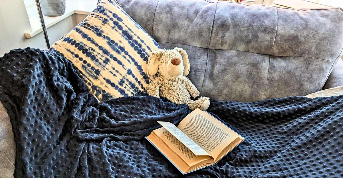 A cozy living room scene featuring a plush gray velvet sofa, a dark navy blue textured blanket, a striped pillow, a small stuffed dog, and an open book.