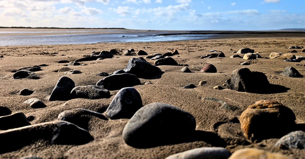 A rocky sandy beach with dark stones in the foreground, calm water, and a blue sky, helping to shape your 2 minute sensory timeout.