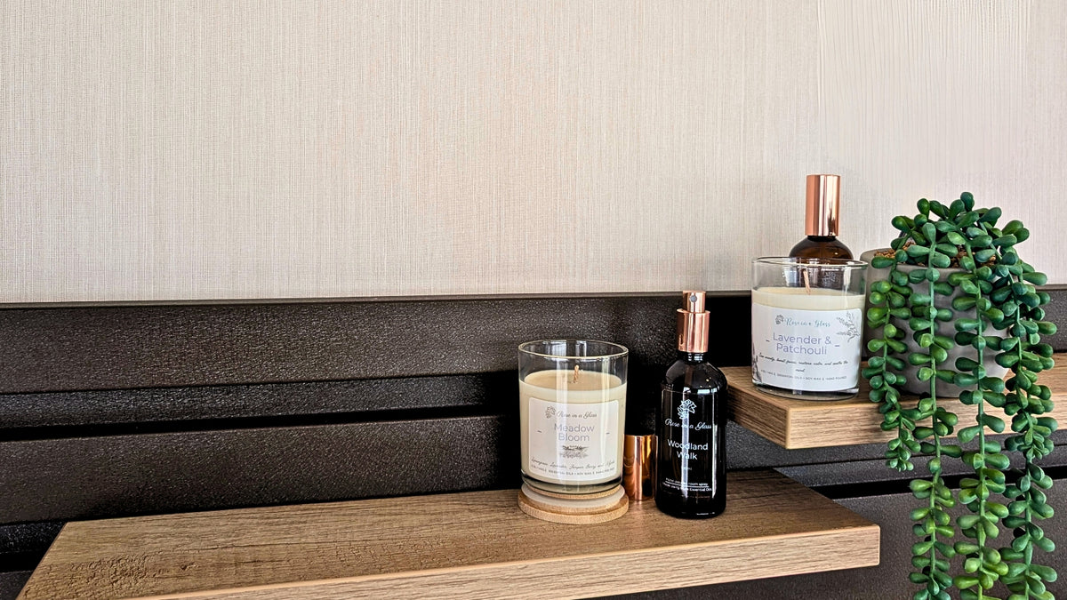 A styled shelf display featuring aromatherapy candles and Room Sprays in reusable glass jars and bottles on a wooden shelf, next to a green trailing plant against a beige textured wall and dark wood paneling.